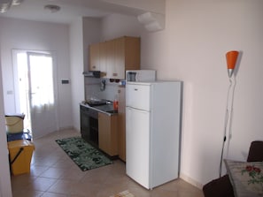 A compact kitchen with a stove, oven, and refrigerator, a small dining area with a table and chair, and a door leading to another room. - SVS - Budoni Sa Capannizza (Budoni)