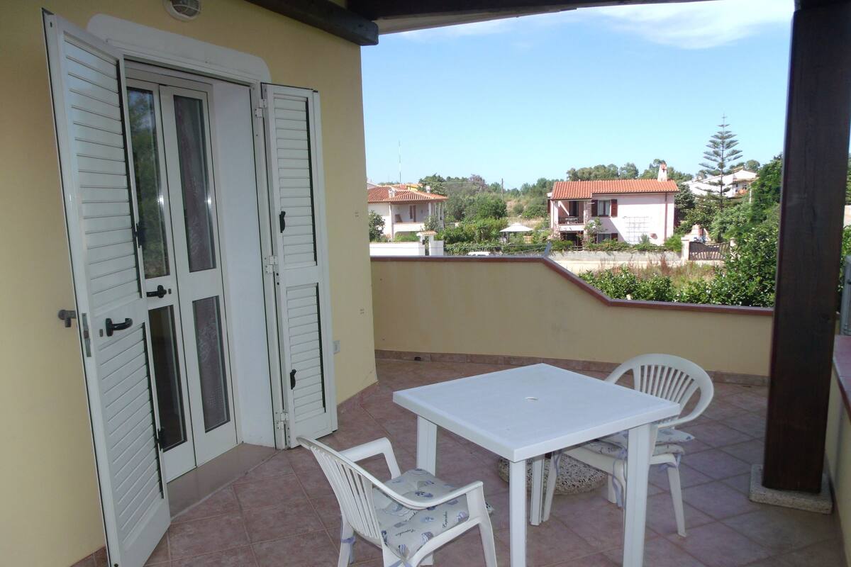 Un balcón con una mesa blanca y dos sillas, un ventanal corredero y vistas a casas al fondo.