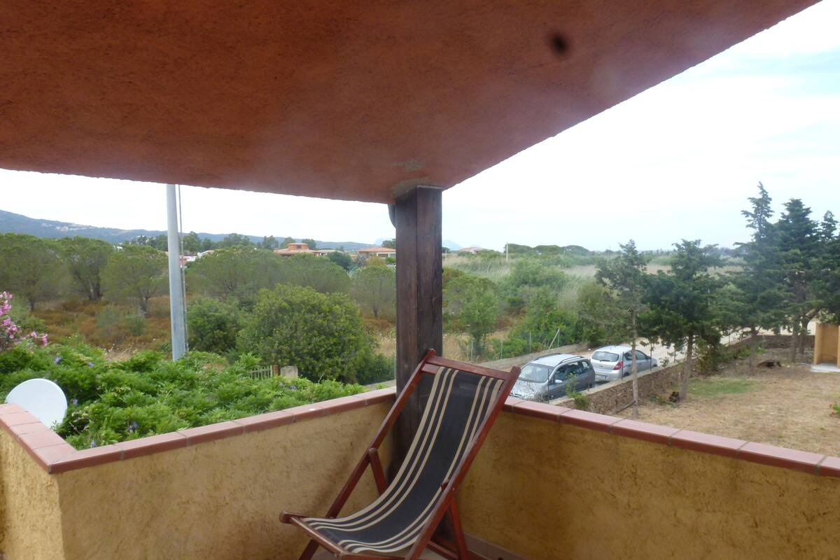 Un balcón con una silla de playa a rayas, vista a un estacionamiento y follaje al fondo.