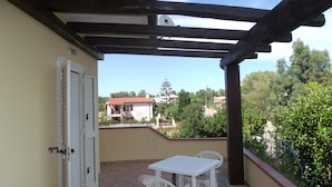 Um alpendre coberto com uma mesa e cadeiras brancas, uma porta e vista para casas e árvores ao fundo.