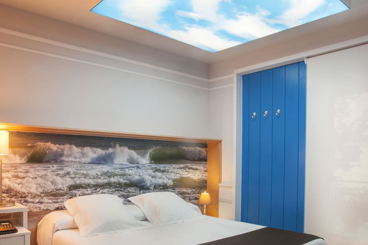 Quarto com cama, mesa de cabeceira, guarda-roupa azul e claraboia.