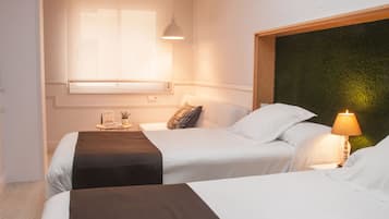 A hotel room with two beds, a skylight, a small table with a lamp, and a window with blinds.