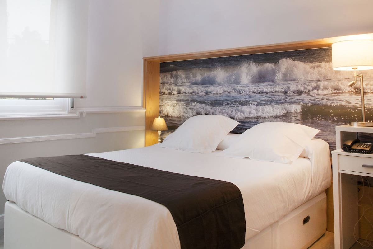 Quarto moderno com claraboia, cama com roupa de cama branca e preta, mesa de cabeceira com abajur e um mural na parede com vista para o oceano.