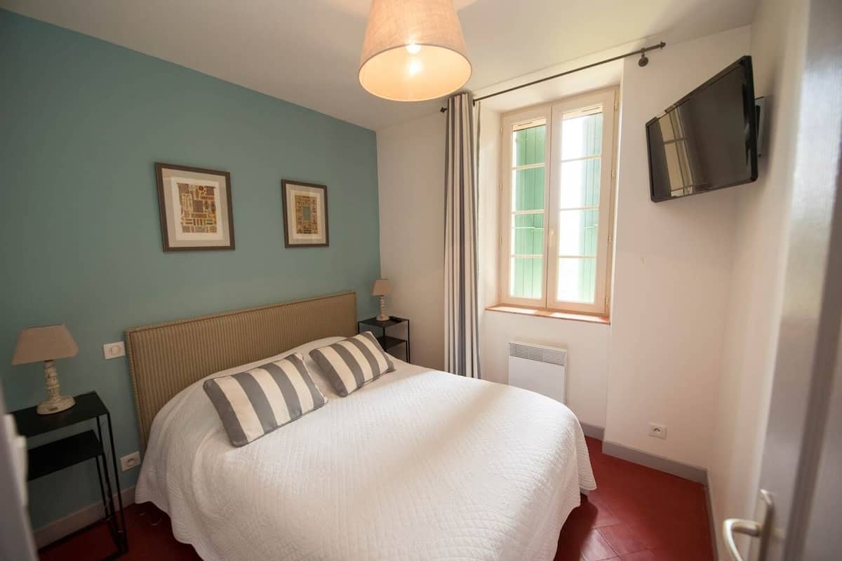 A bedroom with a bed, two framed pictures on the wall, a window with curtains, a radiator, and a TV mounted on the wall.