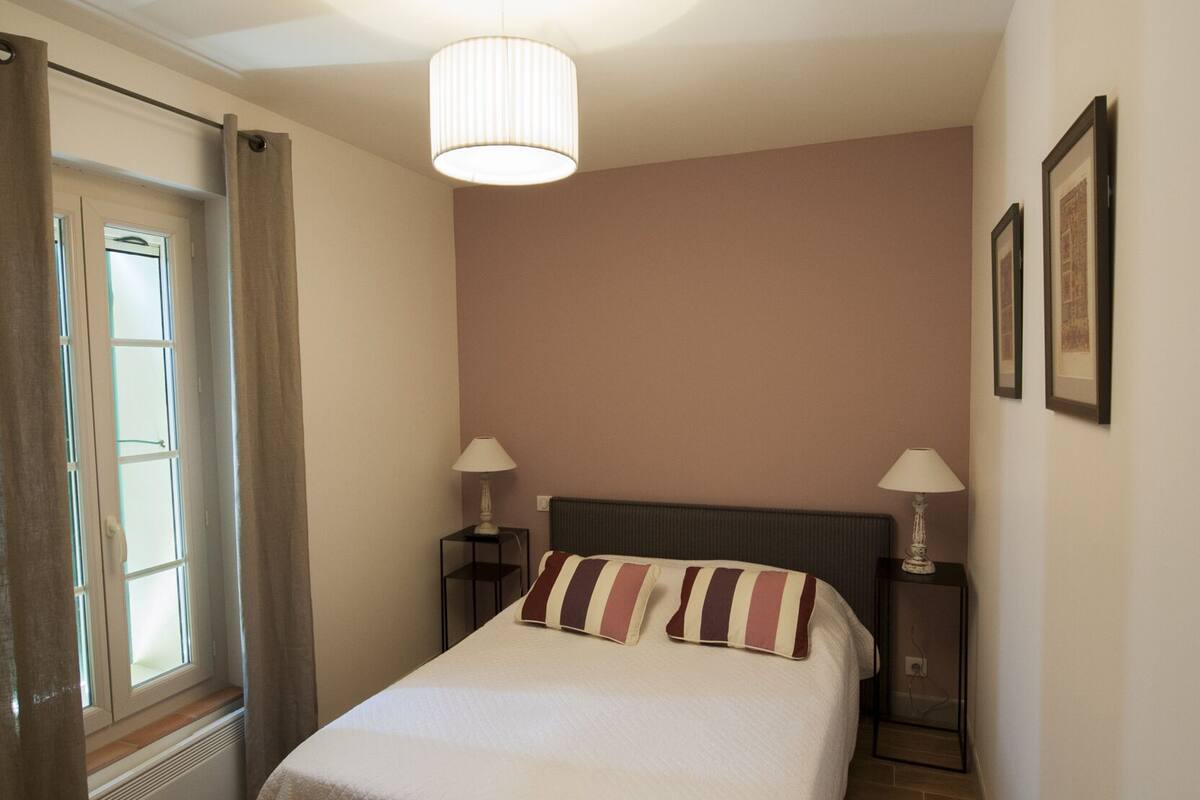 A bedroom with a large bed, two bedside tables with lamps, a ceiling light, and a window with curtains.