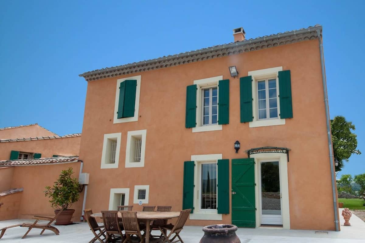 A two-story building with a tiled roof, green shutters, and a central entrance with a door and window.