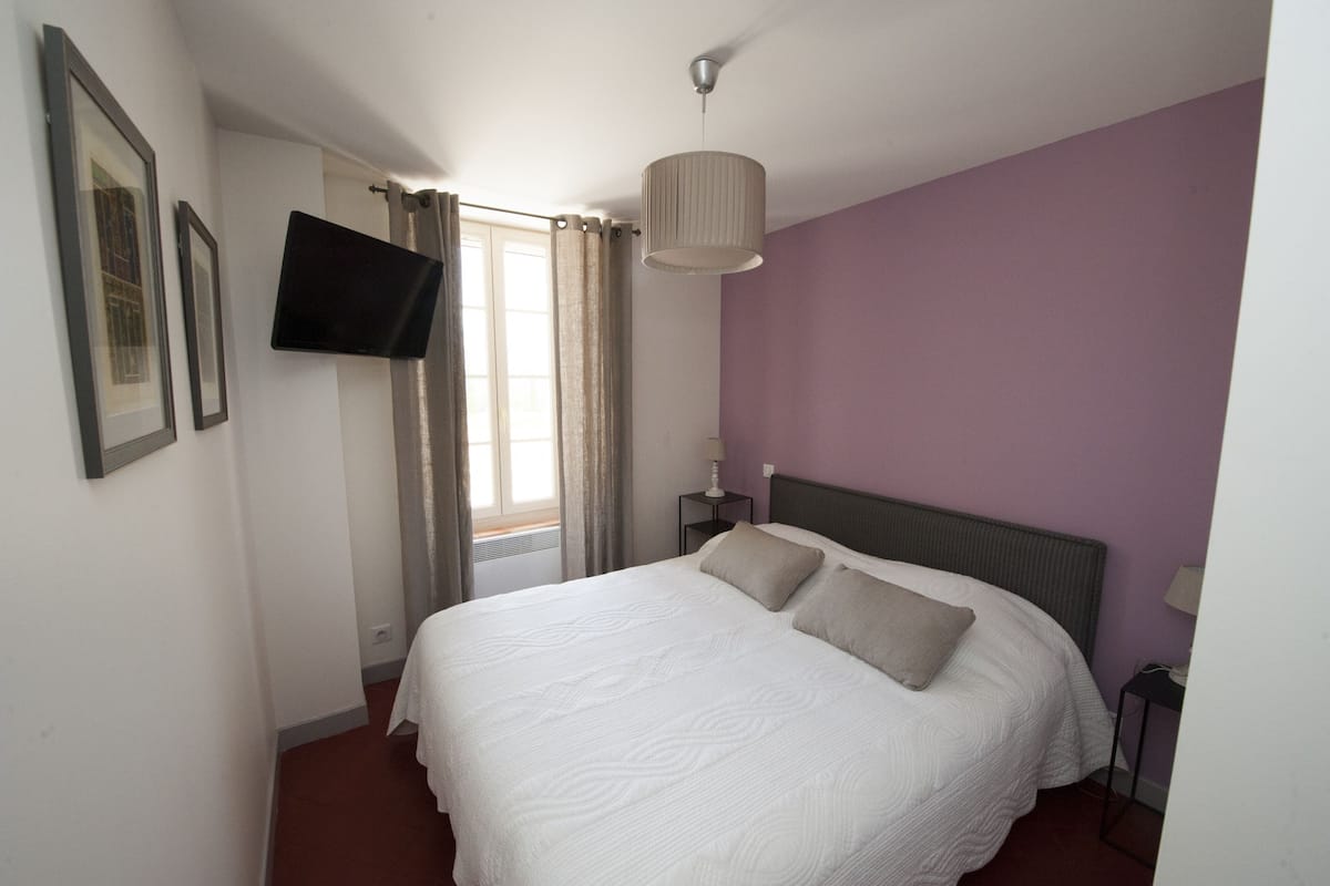 A bedroom with a bed, a television mounted on the wall, a window with curtains, and a bedside table with a lamp.