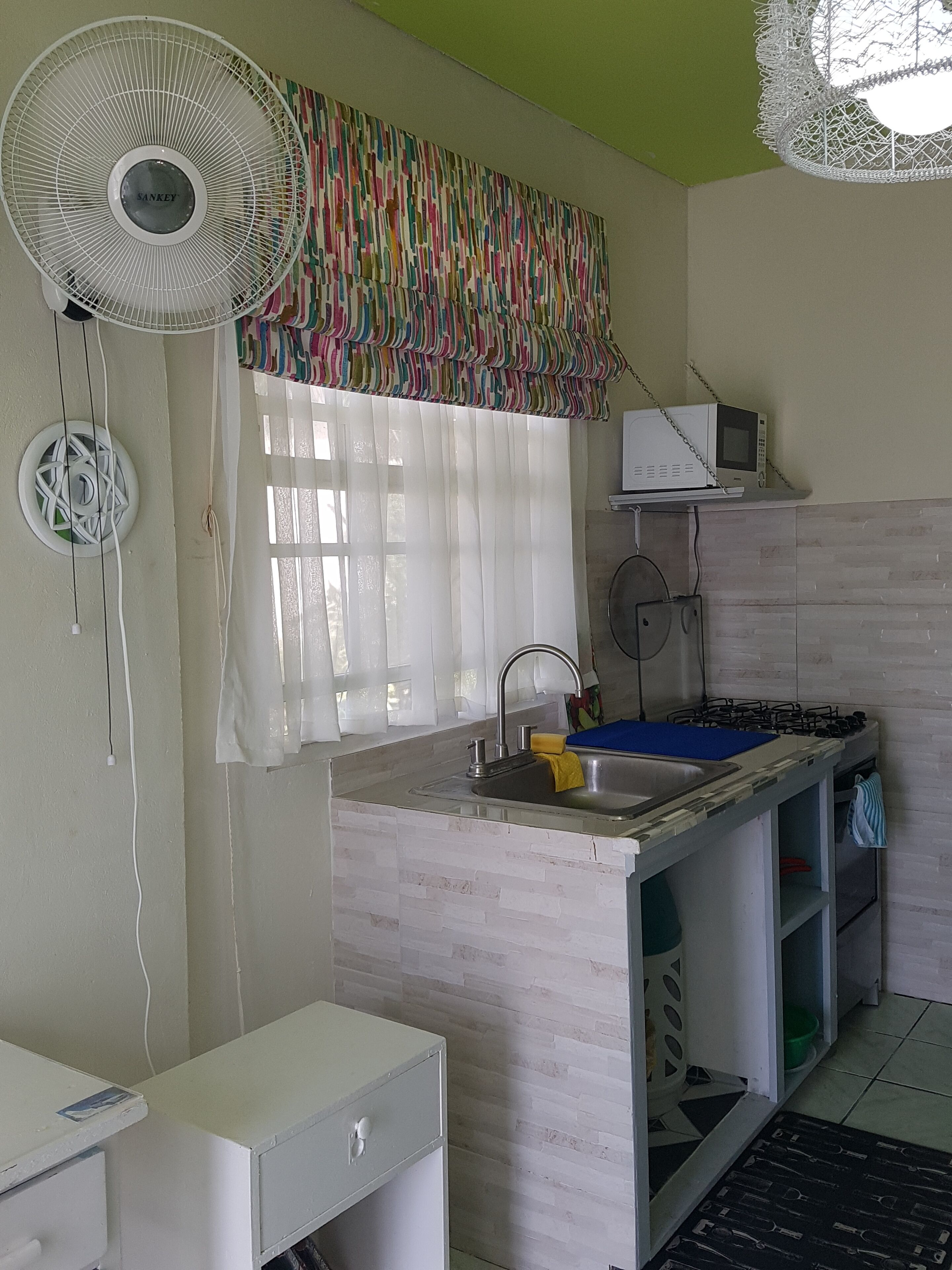 Cocina compacta con un lavamanos, un microondas y una estufa a gas. Hay un ventilador y un mueble pequeño con cajones.