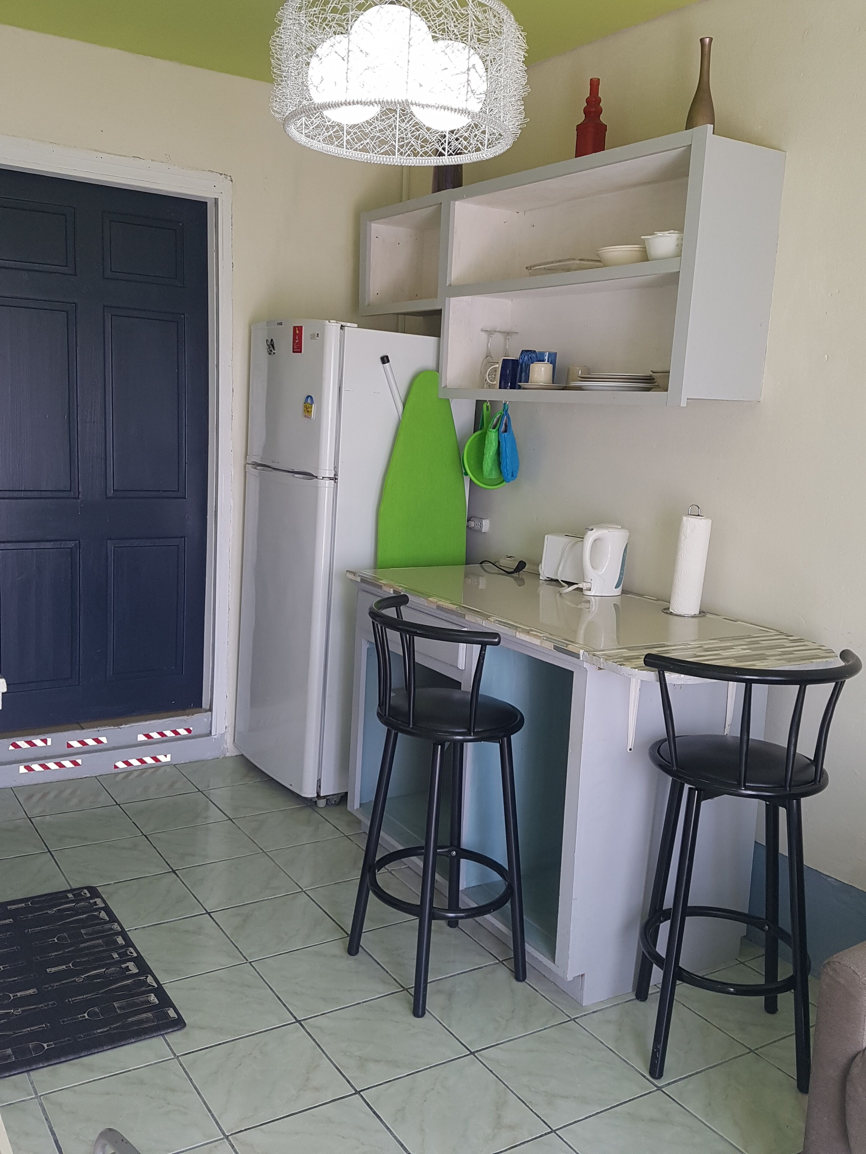 A compact kitchen with a refrigerator, a green chair, two bar stools, and a white countertop.