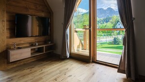 A room with a wooden ceiling, a flat-screen TV mounted on a wooden wall, a wooden floor, and a large sliding glass door leading to an outdoor area with a view of mountains and greenery. - Rezydencja Zakopiańska (Zakopane)