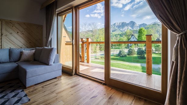 A modern living room with a grey sectional sofa, wooden flooring, and large sliding glass doors leading to a balcony with a mountain view. - Rezydencja Zakopiańska (Zakopane)