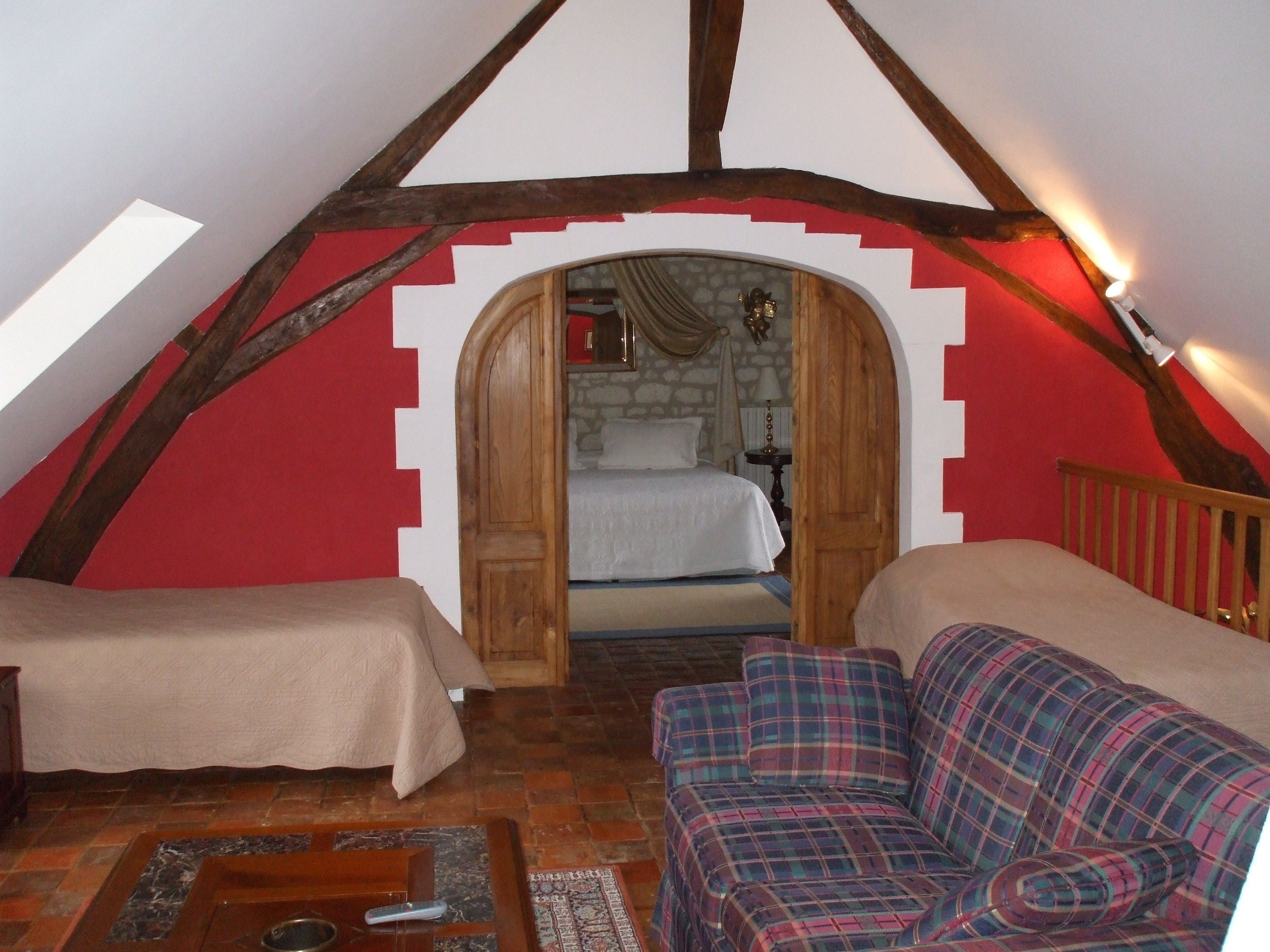 A cozy attic bedroom with a sloped ceiling, a wooden door leading to a bathroom, a bed, a sofa, and a small table with a remote control.