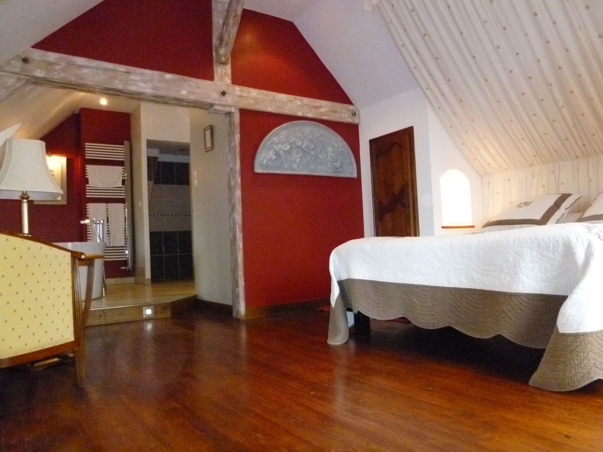 Un dormitorio con piso de madera, una cama con cubrecama blanco, techo de madera y pared roja.