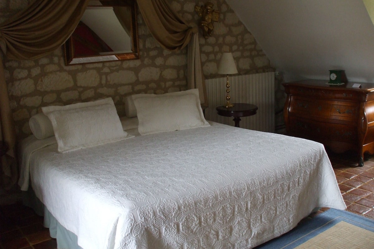 A bedroom with a stone wall, a bed with white linens, a wooden dresser, and a lamp on a small table.