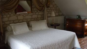 A bedroom with a stone wall, a bed with white linens, a wooden dresser, and a lamp on a small table.