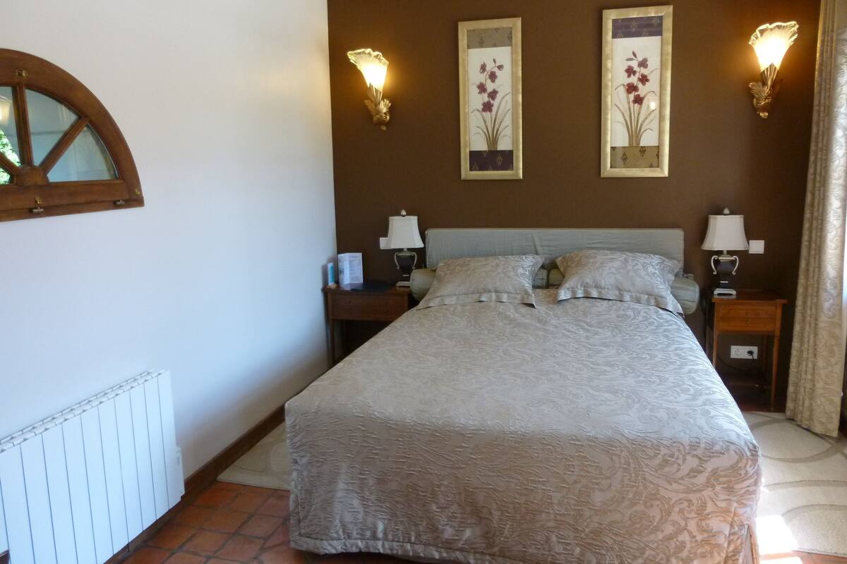 A bedroom with a large bed, two bedside tables with lamps, a window with a wooden frame, and two wall-mounted light fixtures.