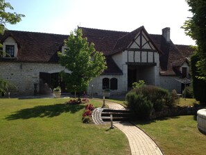 A stone building with a tiled roof, a paved pathway, and a small garden area with a tree and shrubs.