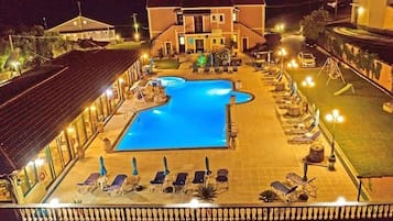 Zona de piscina con sillones de descanso y sombrillas, rodeada de edificios con luces encendidas.