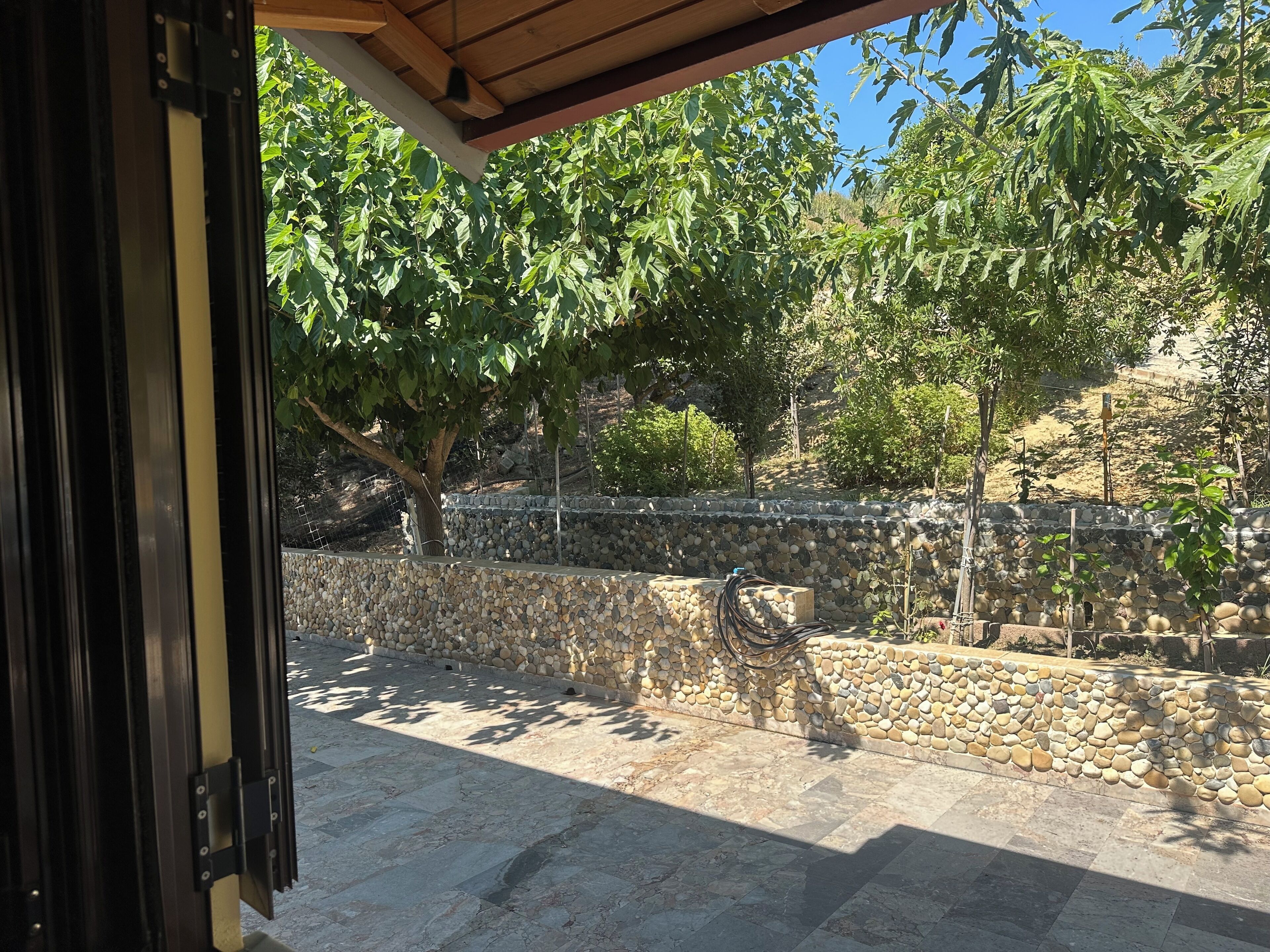 Un patio al aire libre con pared de piedra, pérgola y vista a un jardín con árboles y otra pared de piedra al fondo.