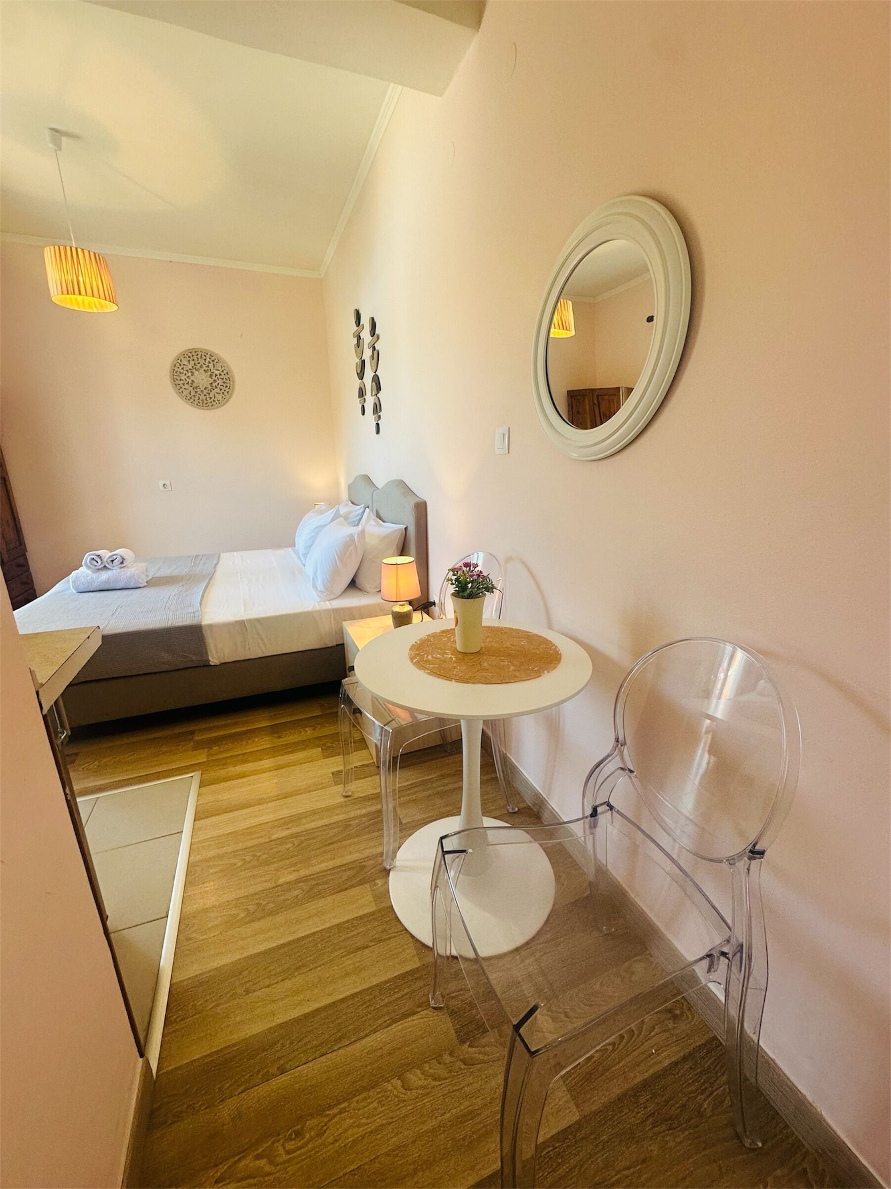 A compact hotel room with a bed, a small table with a lamp, a mirror, and two chairs.
