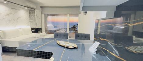 A modern living room with a marble floor, a sofa, a dining table with a placemat, and a view of the sunset through large windows.