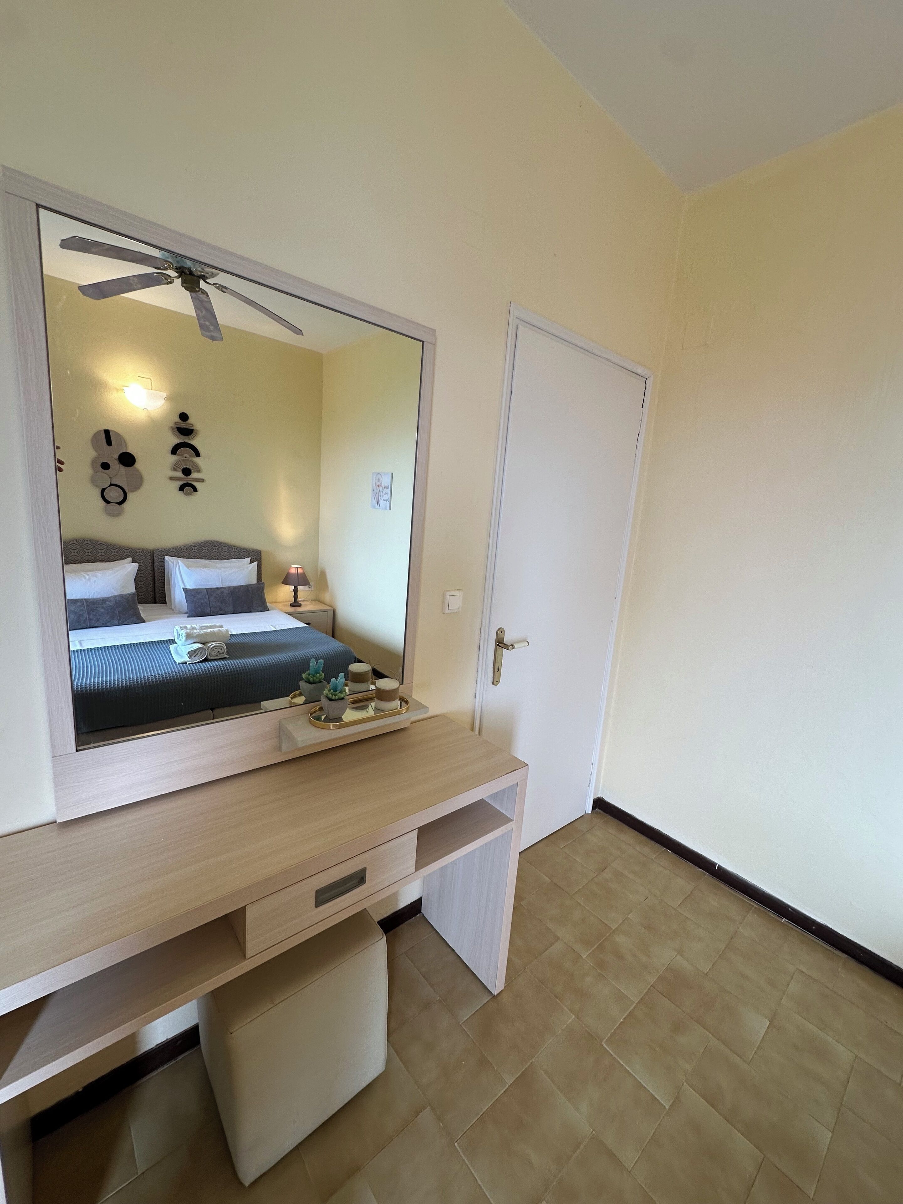 A hotel room with a bed, a desk with drawers, a mirror, a ceiling fan, and a door.
