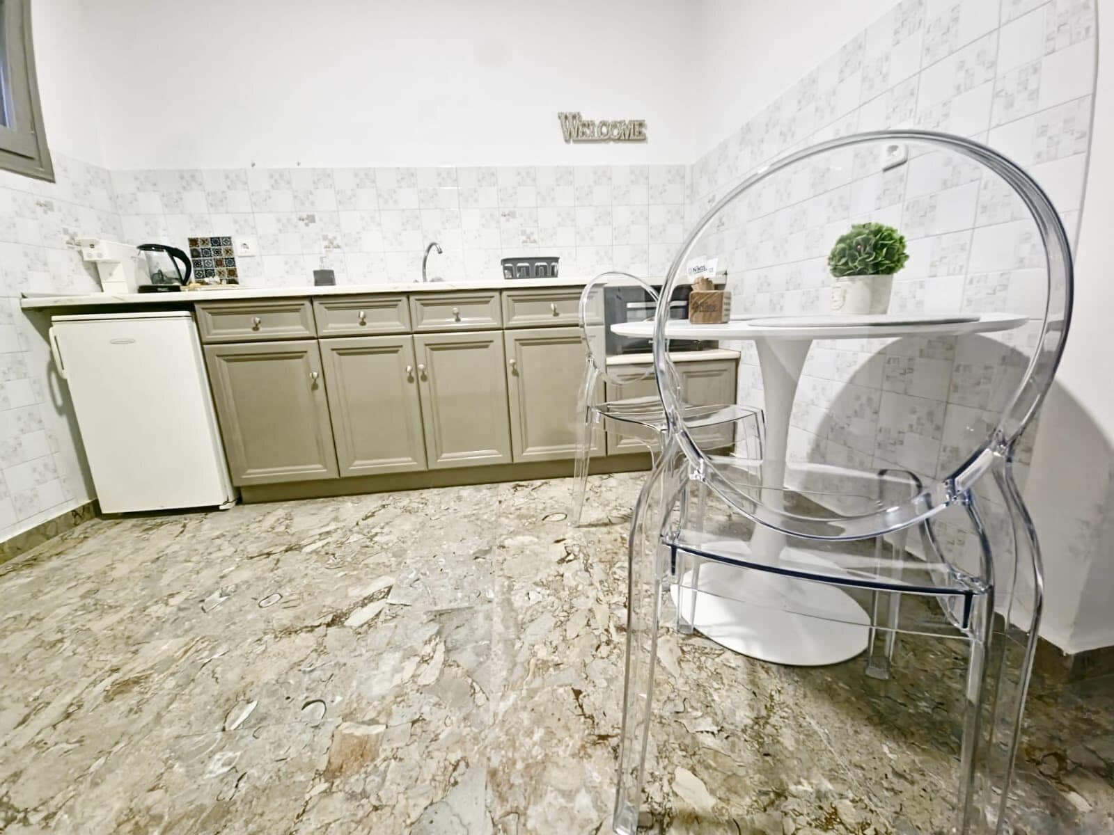 Una cucina con pavimento in marmo, mobili bianchi, un piccolo frigorifero e un cartello di benvenuto.
