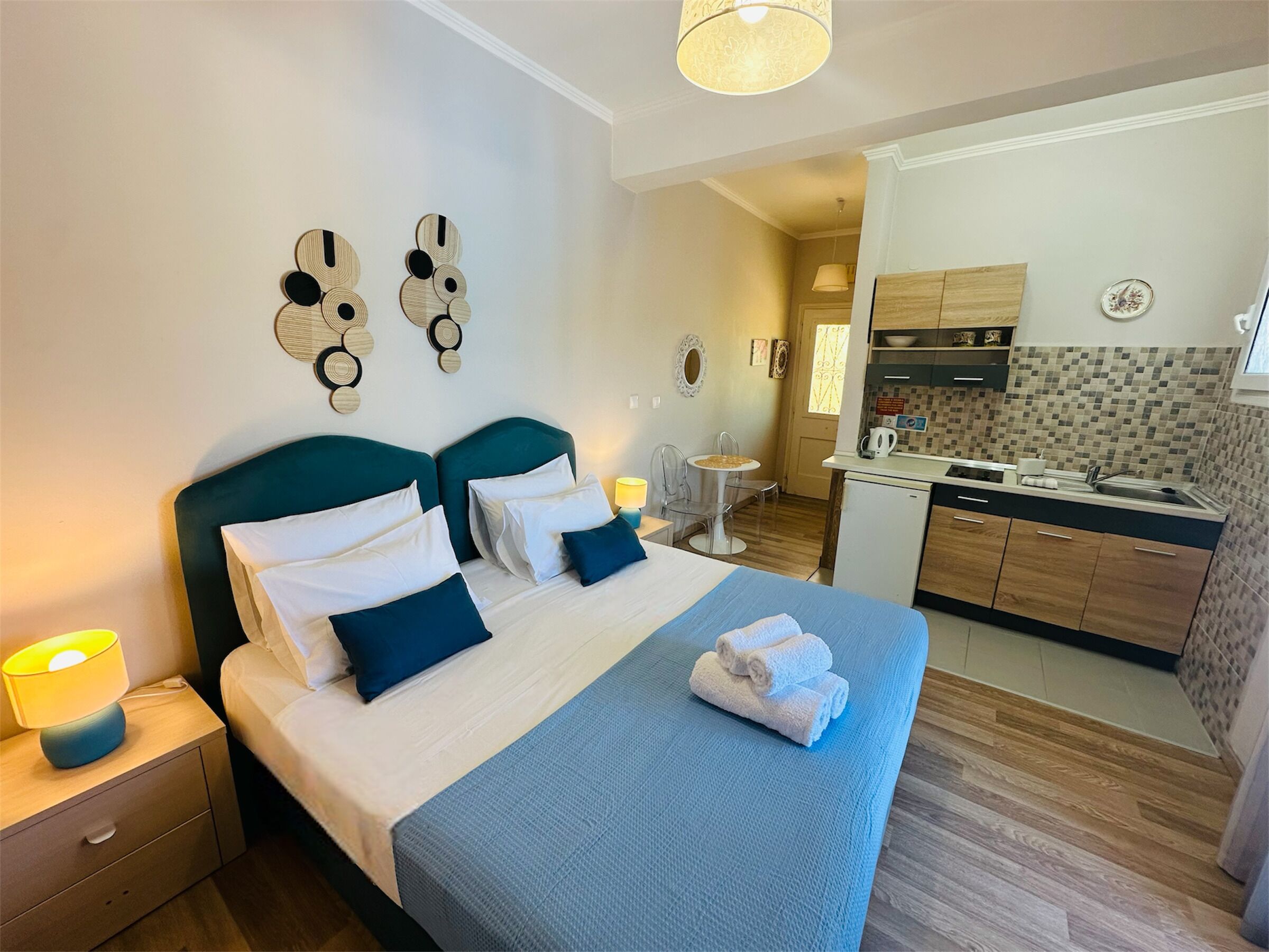A hotel room with a bed, bedside tables, a desk, a chair, a small table, a kitchen area, and a bathroom.