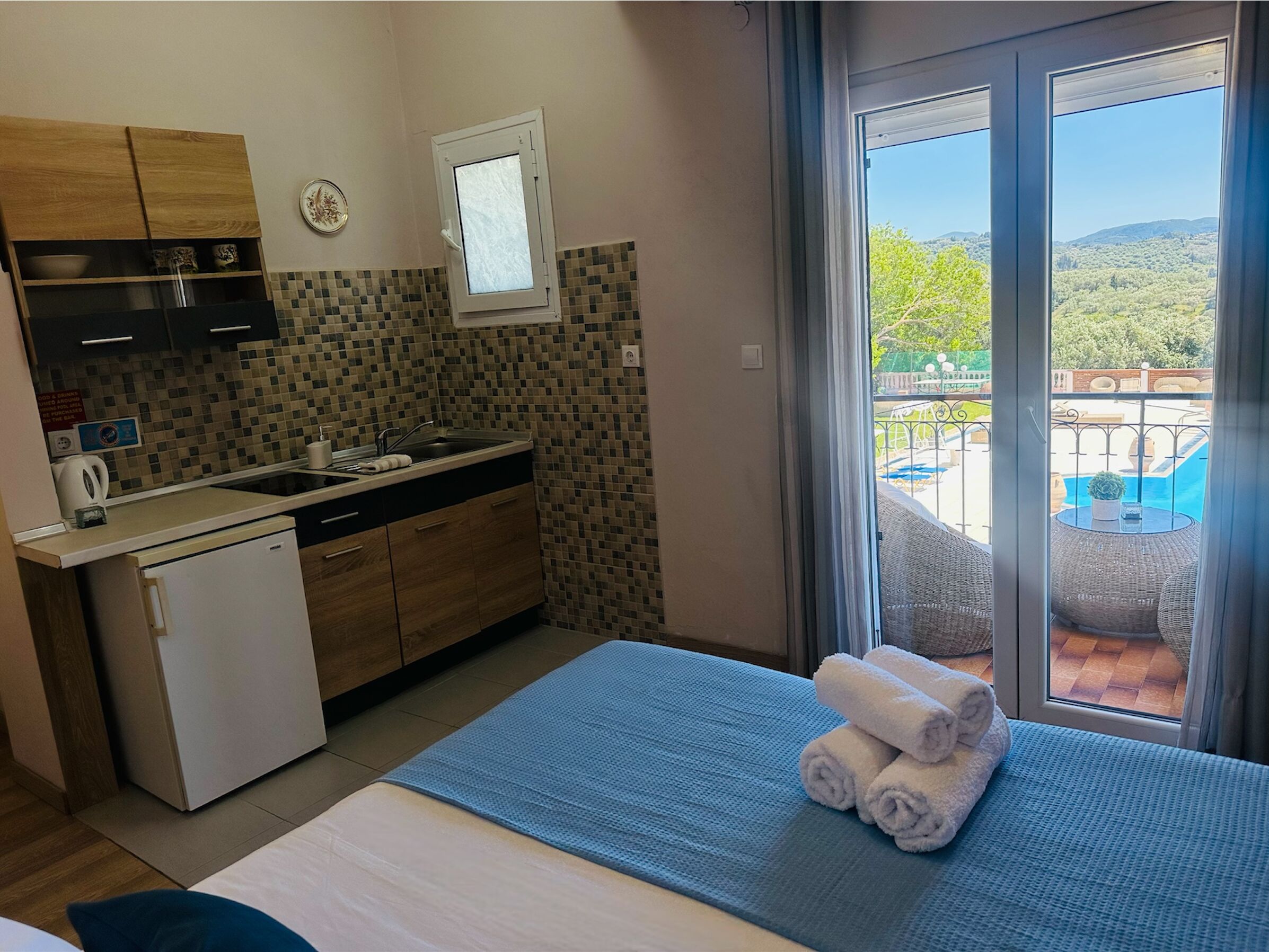 A compact kitchen with a refrigerator, sink, and wooden cabinets. A window offers a view of a pool and greenery.