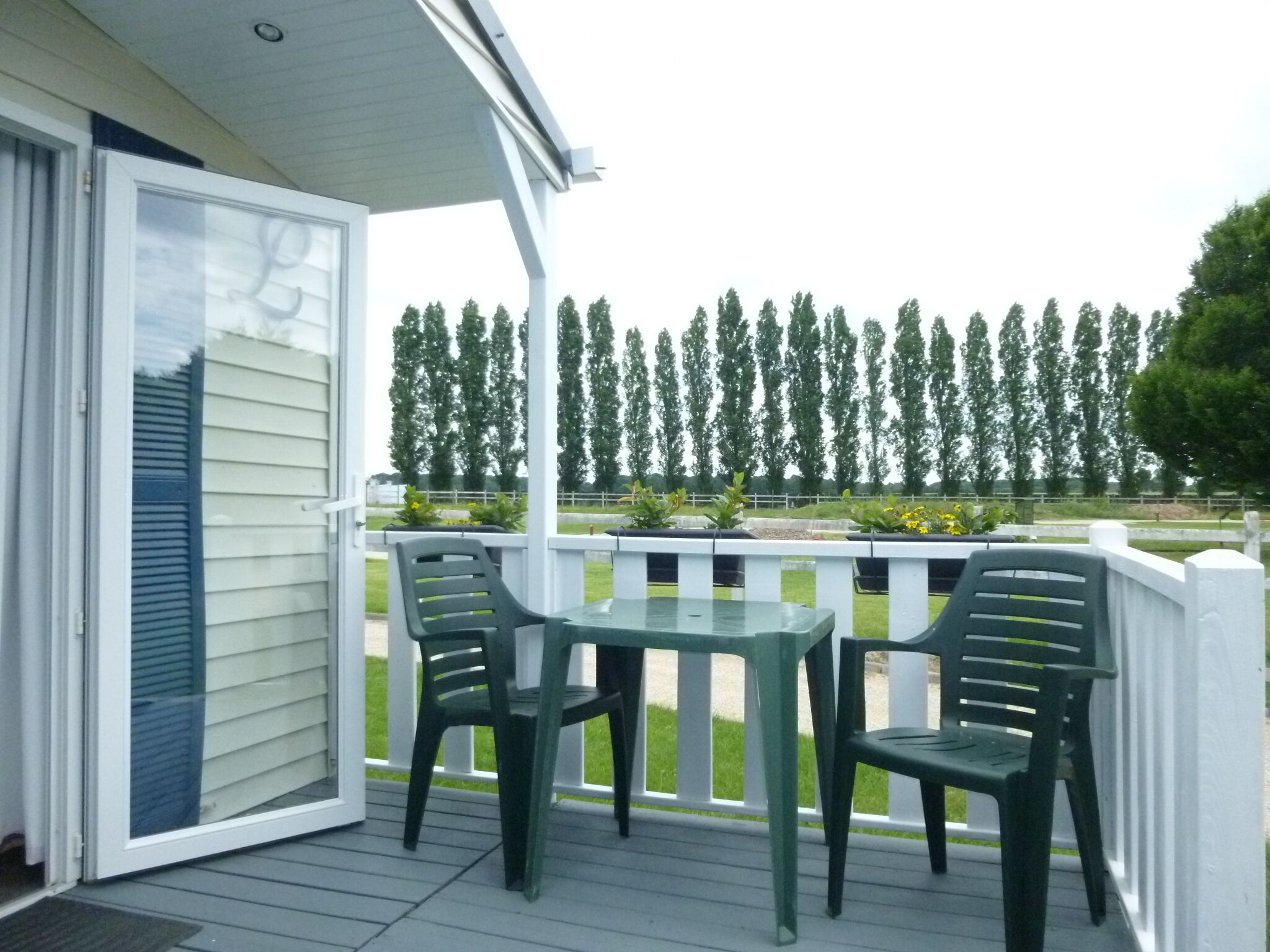 Une terrasse couverte avec une table et deux chaises, donnant sur une rangée d’arbres et une zone herbeuse.