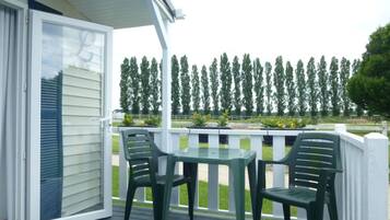 A covered patio with a table and two chairs, overlooking a row of trees and a grassy area.