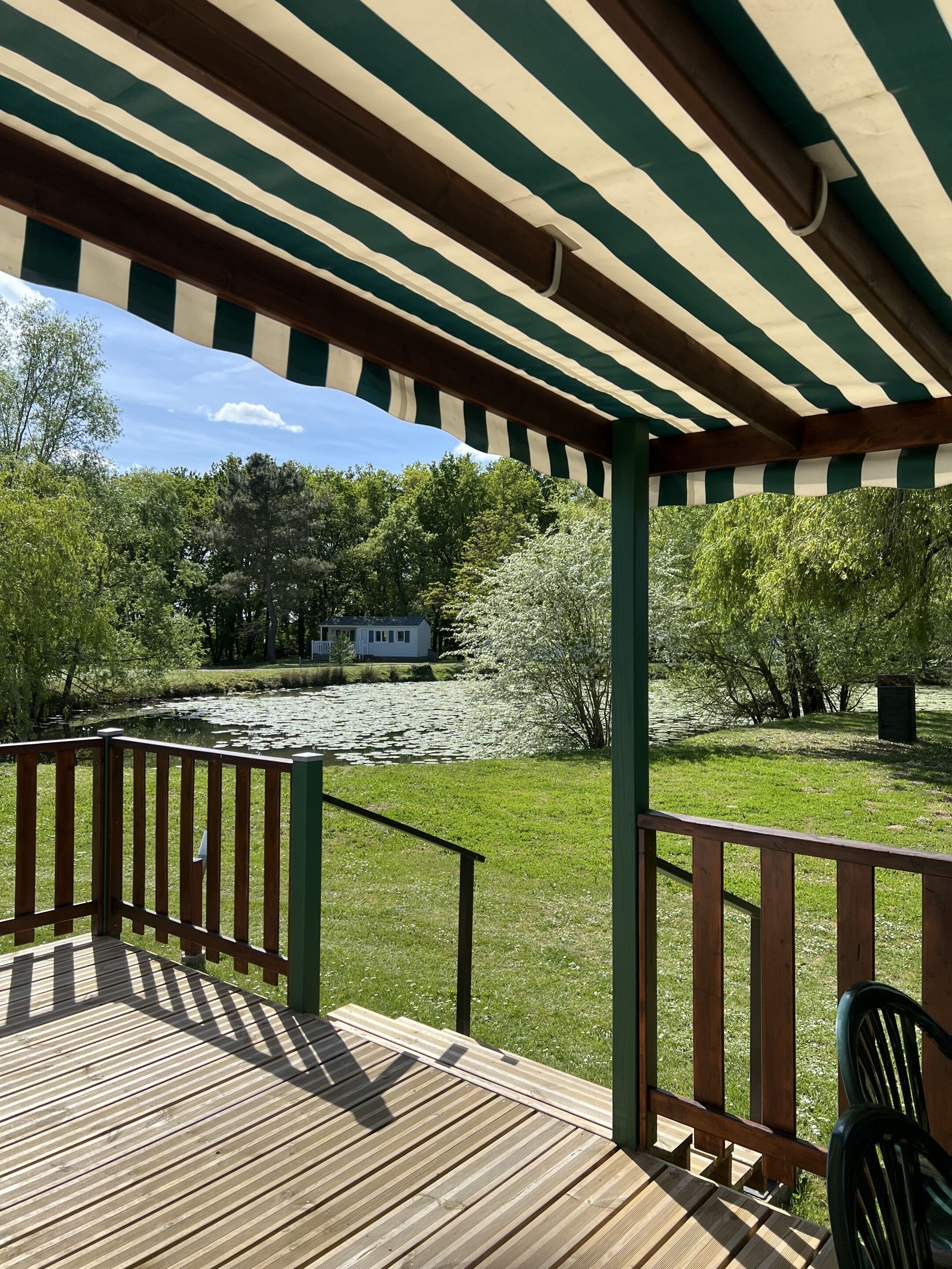 Um deck de madeira com toldo verde e branco às riscas, um lago com vitórias-regias e árvores ao fundo.