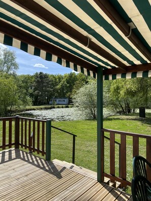 Une terrasse en bois surmontée d’un auvent à rayures vertes et blanches, un étang avec des nénuphars et des arbres en arrière-plan.