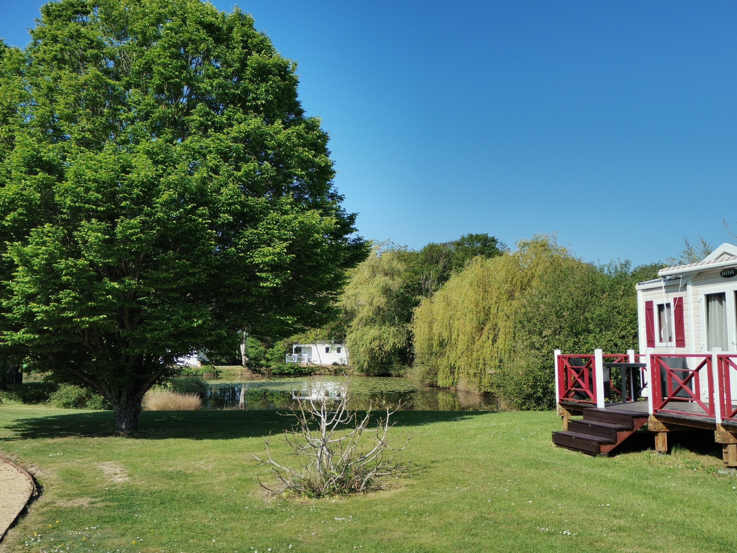 Une maison mobile blanche avec des touches de rouge, une petite terrasse et un étang dans une zone herbeuse bordée d’arbres.