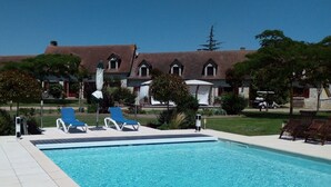 Una piscina con sillones de descanso, un edificio de piedra con techo de tejas, y un cielo azul despejado.