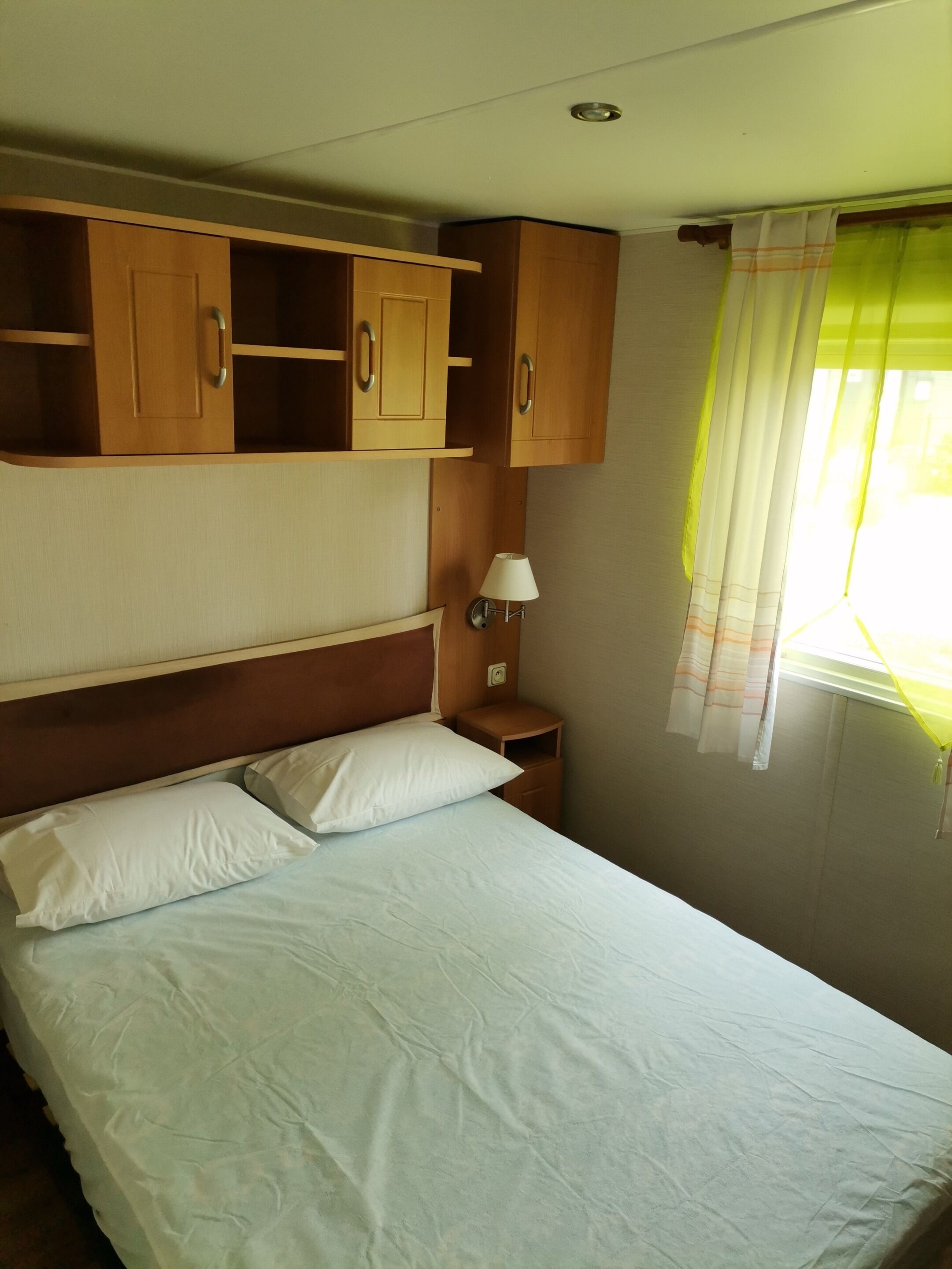 Ein Schlafzimmer mit einem Bett, weißen Kissen, einem Nachttisch, einer Wandleuchte und Holzschränken über dem Bett.