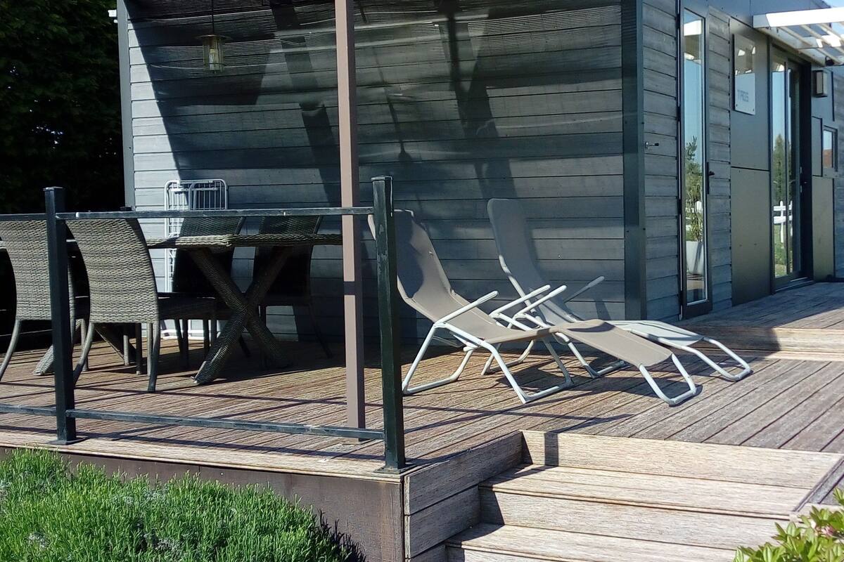 Un espacio al aire libre techado con una mesa, sillas, una terraza de madera y un edificio con amplias ventanas.