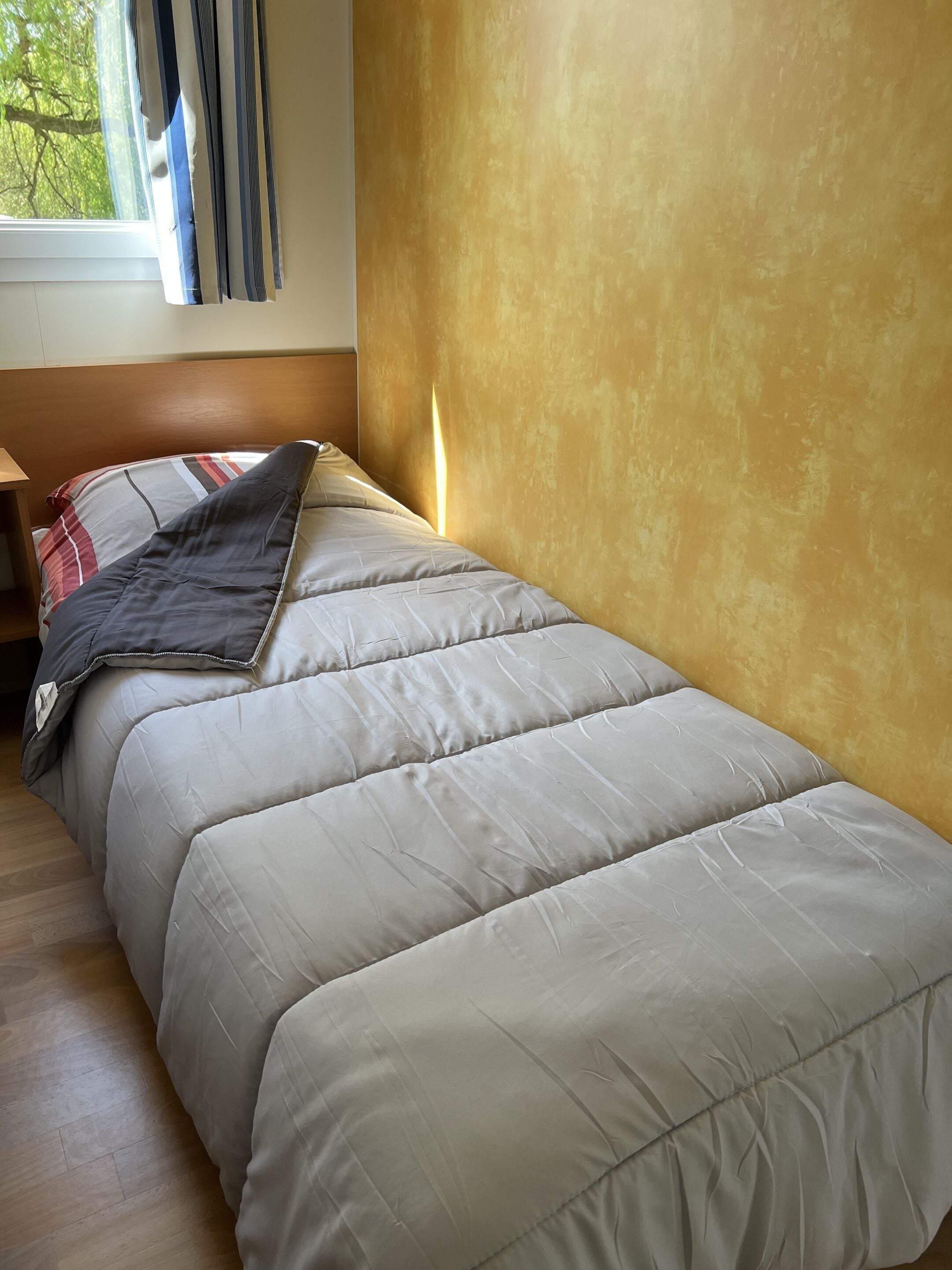 En enkeltseng med et beige dynebetræk, et træhovedgærde, et natbord med en lampe og et vindue med gardiner.