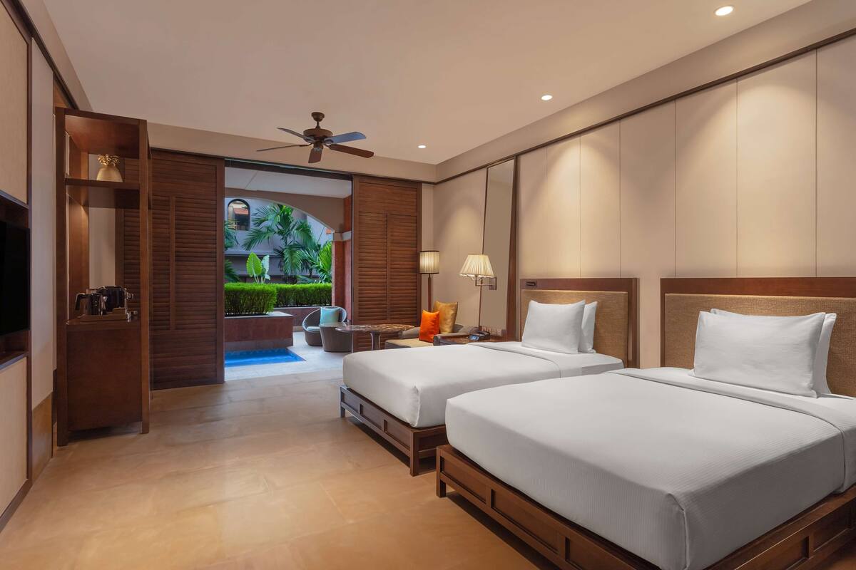 A hotel room with two beds, a ceiling fan, a TV, and a view of a garden.