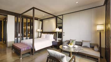 Una habitación de hotel moderna con cama con dosel, una zona de estar con sofá y una pequeña mesa, además de un rincón de descanso con una silla.