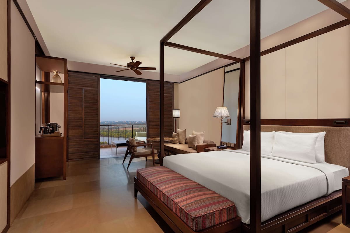 A four-poster bed with a wooden frame, a bedside table with a lamp, a bench with a patterned cushion, and a view of the outdoors through a large window.