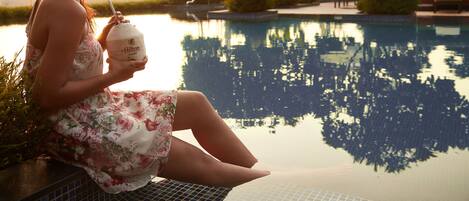 Une femme est assise au bord d’une piscine, un verre à la main, entourée de verdure et bénéficiant d’une vue dégagée sur l’eau où se reflètent les arbres.