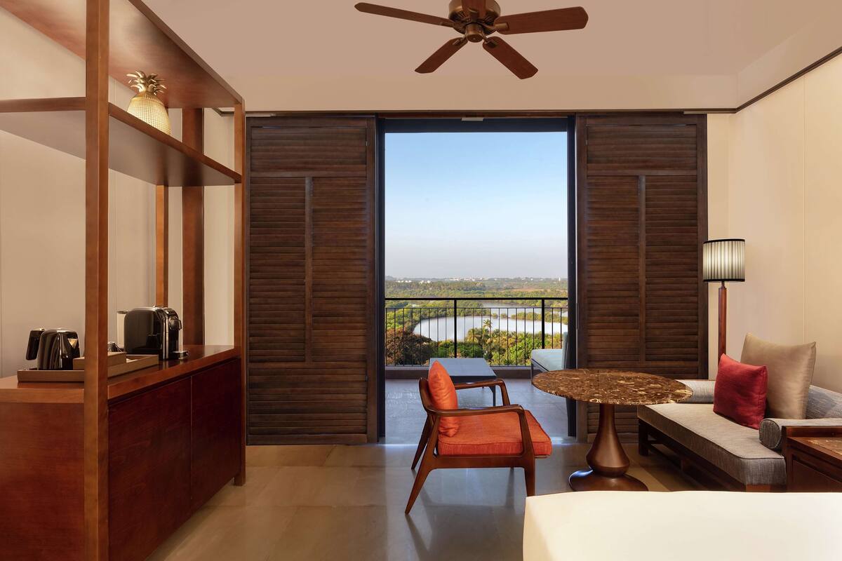 A hotel room with a balcony, a sofa, a chair, a small table, a coffee maker, and a lamp.
