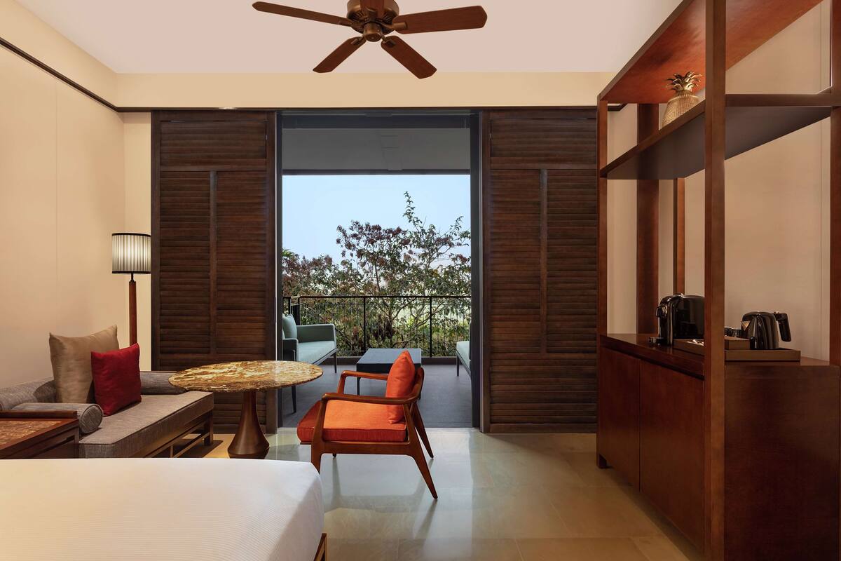 A modern hotel room with a large bed, a seating area with a table, a sofa, and a balcony with a view of trees.
