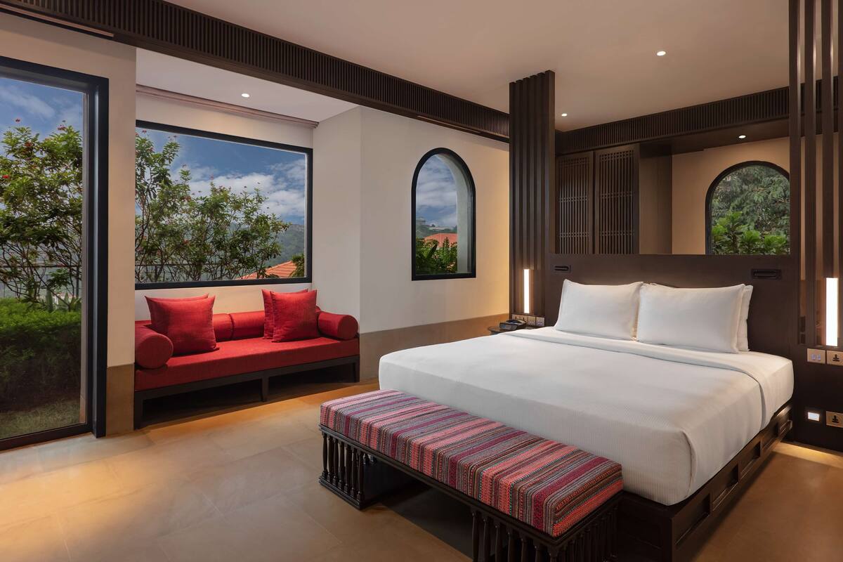 A modern hotel room with a large bed, a red sofa, a bench, and large windows offering a view of greenery and buildings.
