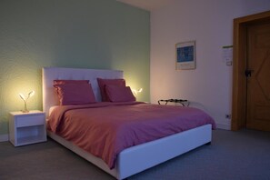 Een net opgemaakt bed met een wit hoofdbord en roze beddengoed, een nachtkastje met een lamp en een houten deur op de achtergrond.