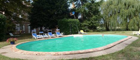 A swimming pool with lounge chairs, surrounded by trees and a building in the background.