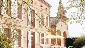 Un edificio de ladrillo grande con una torre, mesas exteriores con sombrillas y un jardín bien cuidado.
