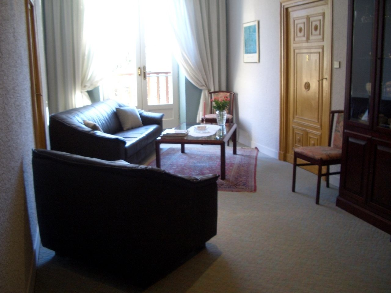 Una sala de estar con un sofá oscuro, una mesa de centro de madera y una silla. Hay una puerta con un número y una ventana con cortinas.