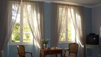 Una habitación con dos ventanas, una mesa con un florero, dos sillas y un televisor sobre un mueble.